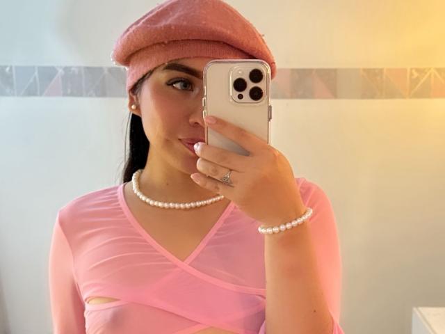ZoeRedds met een roze hoed, een parelketting en een doorschijnend roze topje, waarbij haar borsten zichtbaar zijn, met een telefoon in haar hand, kijkend in een spiegel, met een parelarmband, in een selfie in een badkamer..