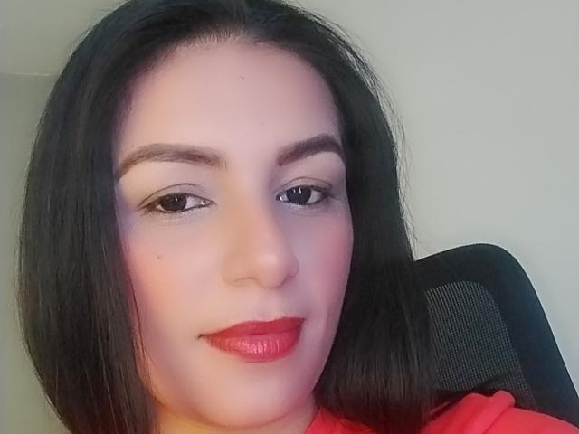 CamilleRiver hat ein hübsches Gesicht mit glattem, schwarzem Haar, rotem Lippenstift und dezentem Augen-Make-up. Sie sitzt auf einem schwarzen Stuhl und lächelt sanft in die Kamera..