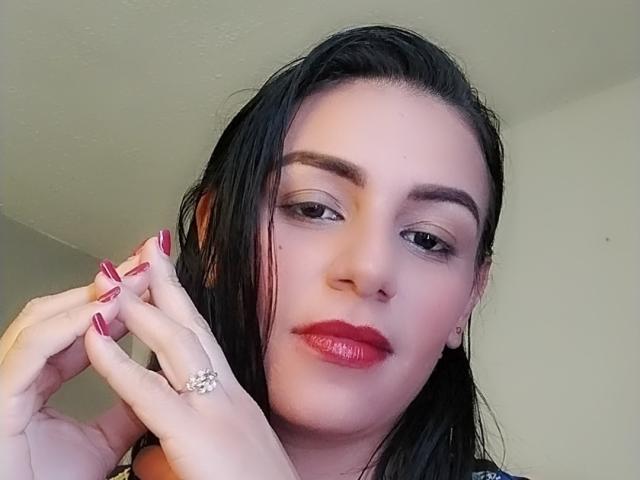 CamilleRiver heeft nat zwart haar, rode lippenstift en rode nagellak. Ze houdt haar vingers dicht bij haar gezicht en laat zo een diamanten ring zien. Haar wenkbrauwen zijn donker en licht gebogen..