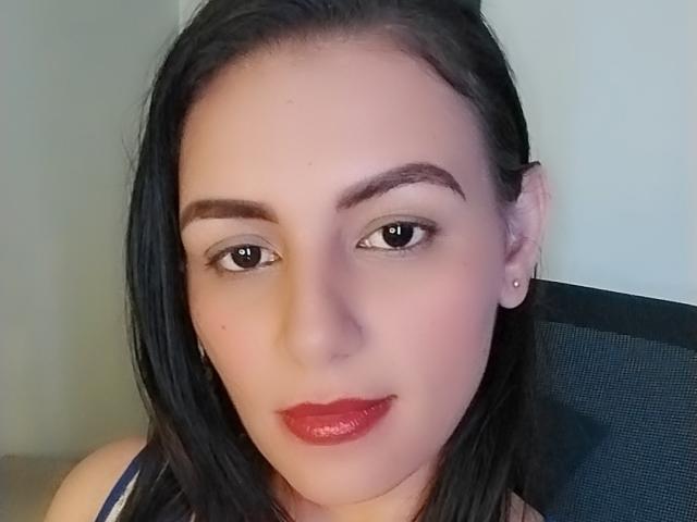 CamilleRiver tiene el pelo negro, piel clara, lápiz labial rojo y cejas oscuras. Lleva una blusa azul y está mirando a la cámara con una ligera sonrisa..
