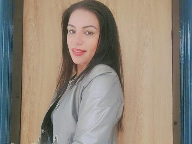 CamilleRiver with long black hair, light skin, and red lipstick, wearing a gray shirt, smiling softly, looking over shoulder, showing a bit of black bra.
