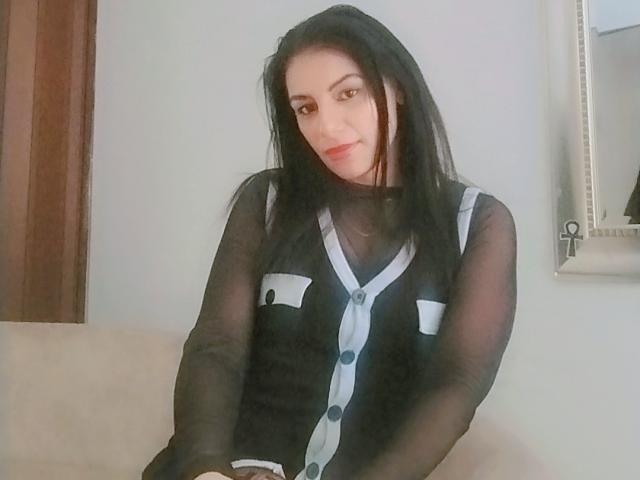 CamilleRiver with long black hair, sheer black top, white buttons, and a slight smile, sitting on a beige couch. She has a cute, attractive face.