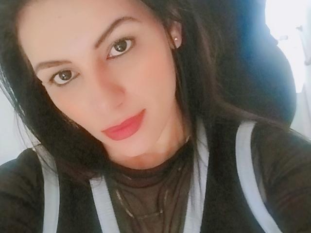 CamilleRiver has pale skin, dark brown eyes, and red lipstick. She's wearing a black shirt with gray straps, black hair, and a small stud earring.
