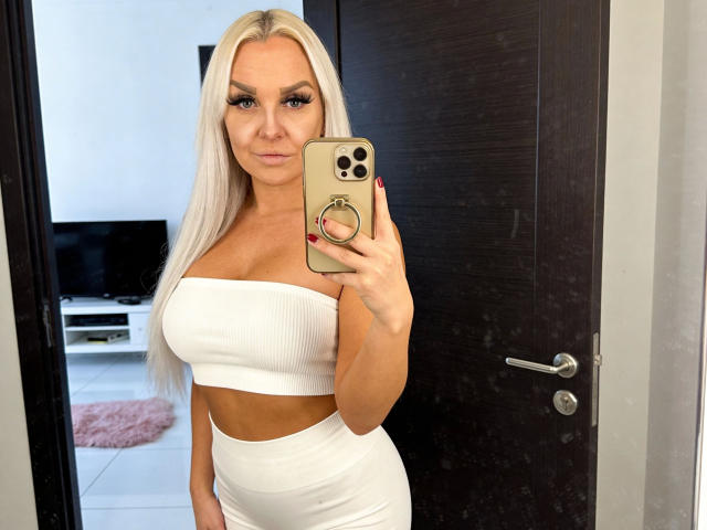 MiaXXX-hot with blonde hair, wearing a white strapless top and high-waisted white shorts, holding a gold iPhone, showing her large tits and curvy ass.