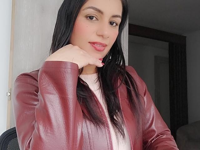 Camille, mit glattem, schwarzem Haar, rotem Lippenstift und heller Haut, trägt eine rote Lederjacke, legt ihr Kinn in die Hand und lächelt leicht in die Kamera..
