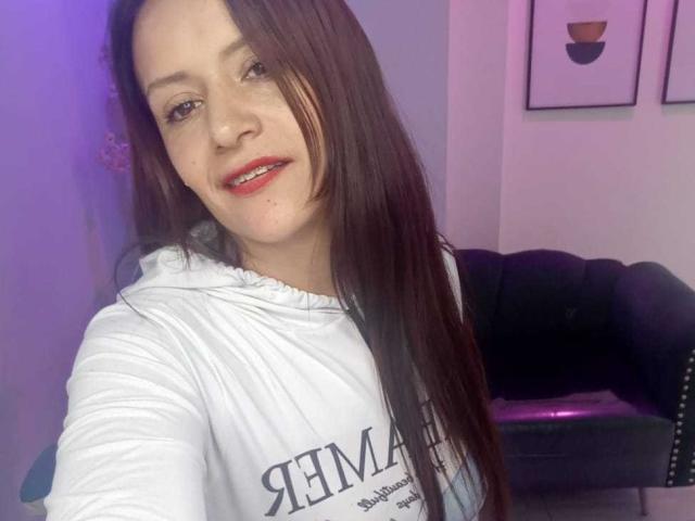 Arianna Seete, com cabelos castanhos longos, moletom branco, batom vermelho, um leve sorriso, pele clara e nariz pequeno, está em pé em um quarto com iluminação roxa. Os seios não estão visíveis..