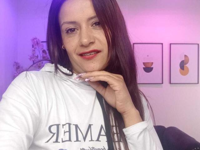 Arianna Seete, com cabelos castanhos longos, batom vermelho, moletom branco e um anel no dedo, sorrindo para a câmera. Iluminação rosa e roxa. Arte na parede..