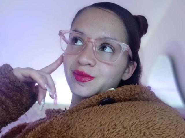 LesleyGray with pink glasses, red lipstick, and brown hair in a bun, holding a brown fuzzy teddy bear, looking up with a cute smile. Her nails are long and white.