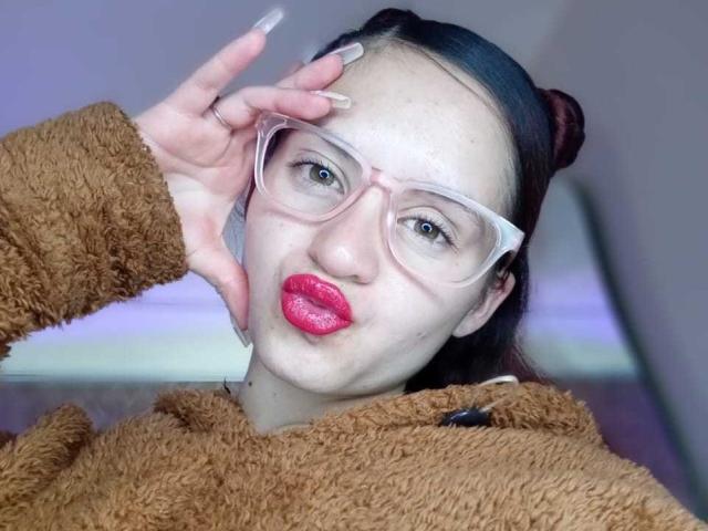 LesleyGray with pale skin, red lipstick, and glasses, wearing a brown fuzzy jacket, has dark hair in buns, and is making a peace sign with long, manicured nails.