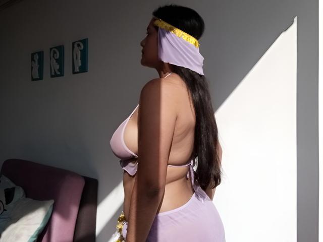 ArianithaXox con cabello largo y oscuro, top de bikini púrpura que muestra un busto grande, y parte de abajo púrpura a juego, pañuelo amarillo en la cabeza, de pie de lado, la luz del sol proyectando una sombra, pared blanca, cama púrpura en el fondo..