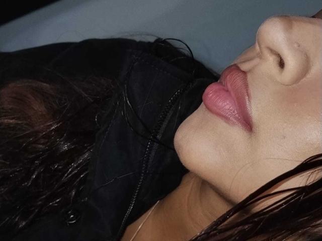 O rosto de BonnieBodn, com lábios rosados e brilhantes, levemente entreabertos, vestindo uma jaqueta preta, com cabelo escuro e molhado, em close-up, mostrando apenas o nariz até o lábio inferior..