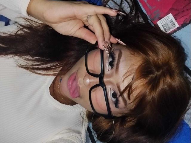 BonnieBodn with brown hair, glasses, pink lips, white top, and long nails, lying down, touching glasses with right hand, looking at camera, showing a cute, sexy face.