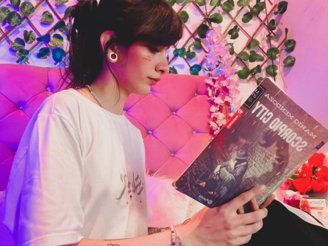 LaurenEvie69 with brown hair in a bun, white shirt, holding "Wardringer" book, purple and pink lighting, earbuds in, pink tufted headboard, green vines, focused on book.