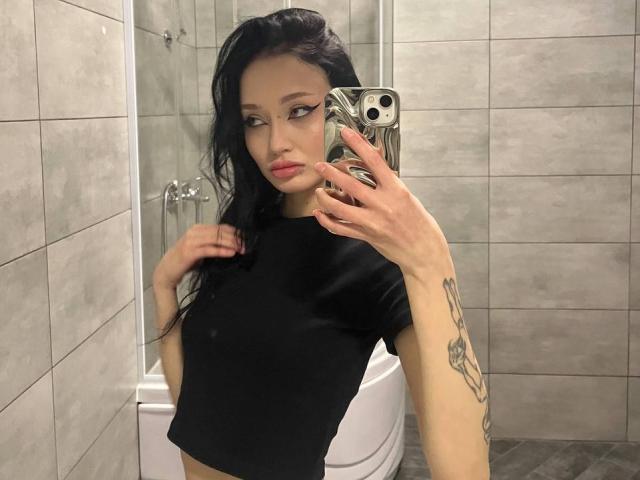 SakuraBloom with long black hair, wearing a tight black crop top, holding a phone with a patterned case, showing a tattoo on her right arm, in a bathroom.