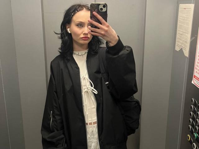 SakuraBloom has black hair, white earbuds, and a black jacket over a white dress with a ribbon. She's taking a mirror selfie in an elevator.