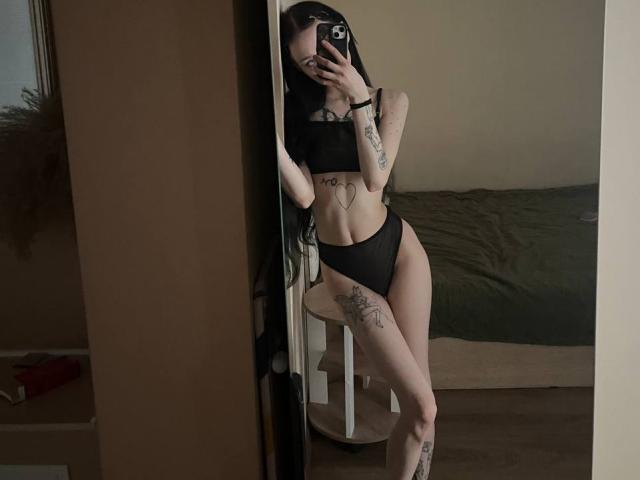 SakuraBloom with black bra and panties, tattoos on arms, legs, and side, holding phone, standing in mirror, long black hair, slim with small tits, in dimly lit room.