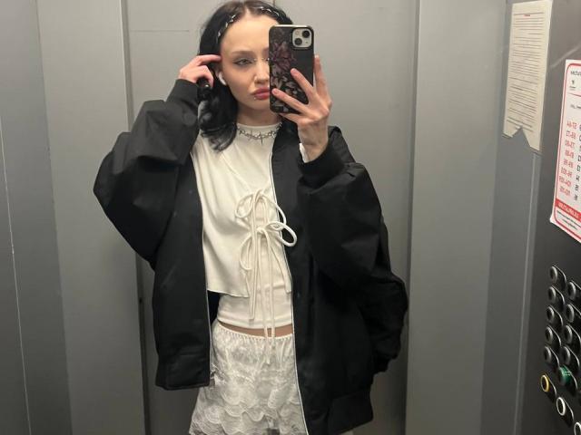 SakuraBloom with black jacket, white lace dress, and necklace, taking mirror selfie in elevator, showing off small tits and slim figure, holding phone with dark case.
