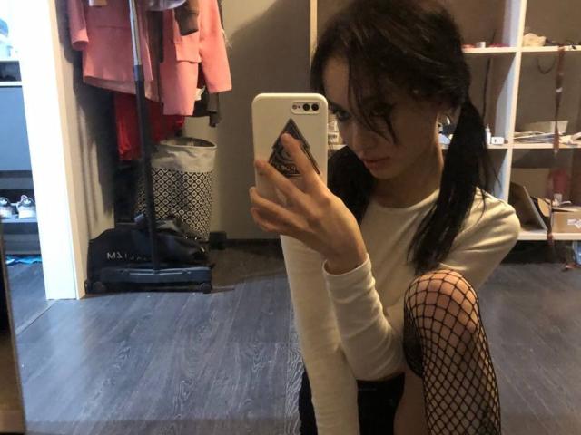 SakuraBloom with pigtails, white long-sleeve top, fishnet leg, squatting, taking selfie in mirror, holding white phone, showing ass, in dimly lit room, wearing earrings.
