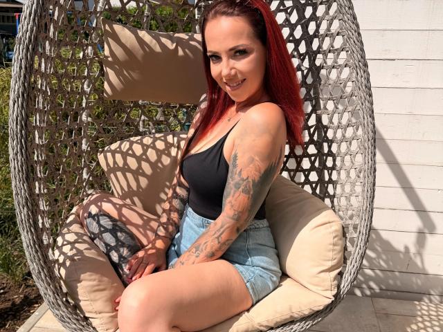 NinaDevil, met rood haar, een zwart tanktopje en denim shorts, zittend in een geweven hangstoel, lachend, met tatoeages zichtbaar op haar rechterarm en linker dij..