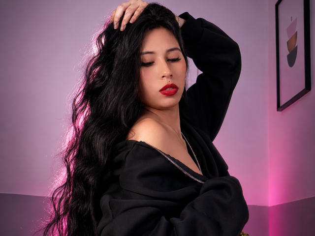 LilaaGrace with long, wavy black hair, red lipstick, and closed eyes, wearing a black off-shoulder top, touching her head, pink and purple lighting, looking sexy.