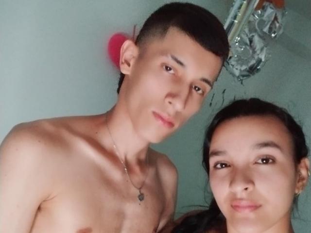 AlannaAndJames, shirtless with a slim build, short brown hair, and a necklace, stands next to a smiling woman with dark hair. Both have fair skin and small, cute tits.