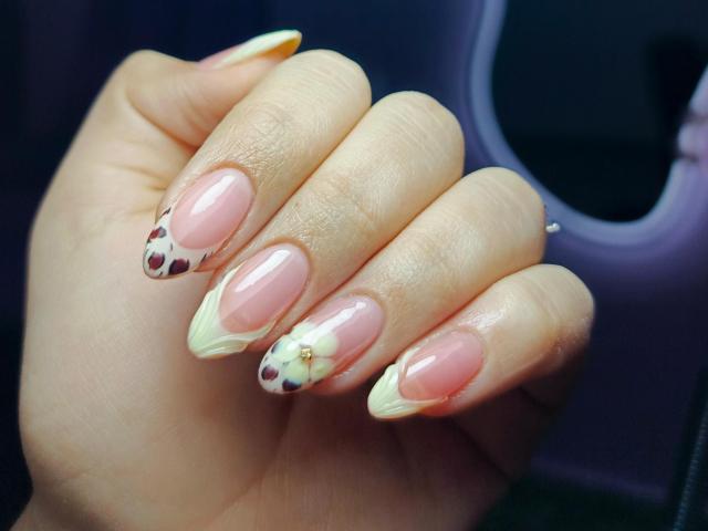 A mão de LissyCarter, com unhas rosa brilhantes, pontas finas e detalhes brancos. As unhas têm pontos pretos e pequenos detalhes decorativos dourados. Sua pele é clara. O dedo está ligeiramente curvado..