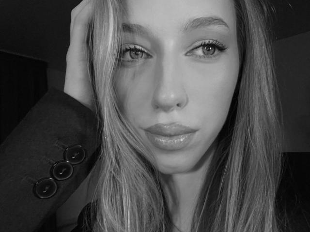DionHolster hat glänzende Lippen, lange Wimpern und einen ernsten Gesichtsausdruck. Sie berührt ihr blondes Haar mit einer Hand und trägt eine dunkle Jacke mit Knöpfen, in Schwarz-Weiß..