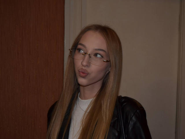 DionHolster with long blonde hair, wearing glasses, black leather jacket, and white shirt, looking to the side with pouty lips.