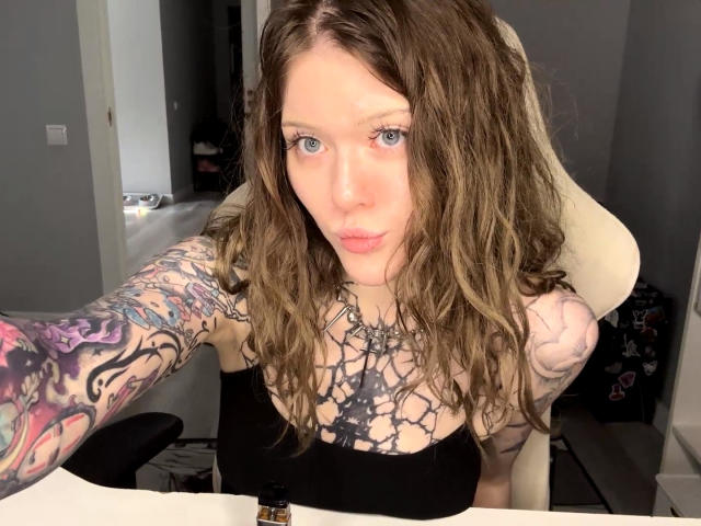 MeliaRinaldo with wavy brown hair, blue eyes, black mesh top, and colorful tattoos on her right arm, taking a selfie in a white chair, with a neutral expression.