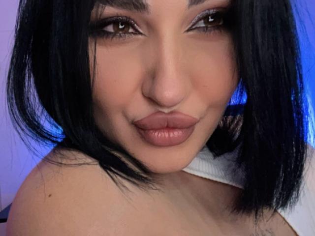 BoobZillaa ha gli occhi marrone chiaro, labbra piene rosa e capelli neri lunghi fino alle spalle, con un leggero sorriso. La sua pelle è marrone chiaro e indossa un reggiseno con spalline bianche..