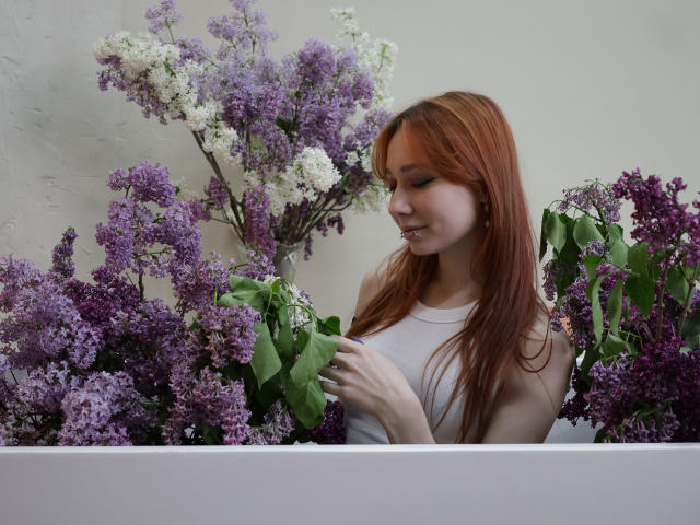 AdrianeWalter with red hair, small tits, wearing a white tank top, surrounded by purple and white lilacs, holding green leaves, smiling, in a white box.