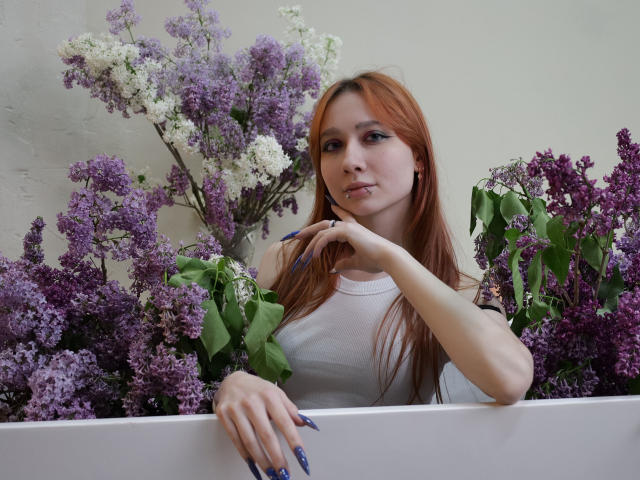 AdrianeWalter with red hair, white tank top, and purple nails, leaning on a white surface surrounded by purple and white lilacs. Her hand touches her chin.