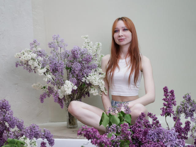 AdrianeWalter, red-haired, sitting cross-legged, wearing a white crop top, surrounded by purple and white lilacs, smiling softly, pale skin, small tits, and a cute, innocent face.