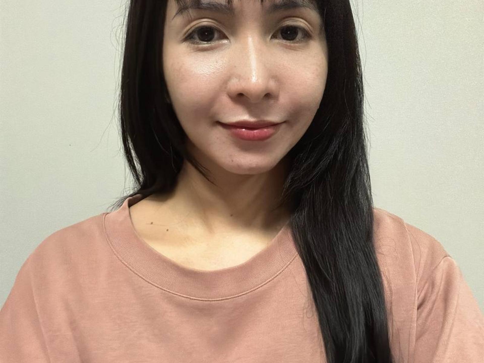 DeaneWilhite with straight black hair, light skin, small nose, pink shirt, subtle smile, and pink lips, showing a clean, soft-faced look.