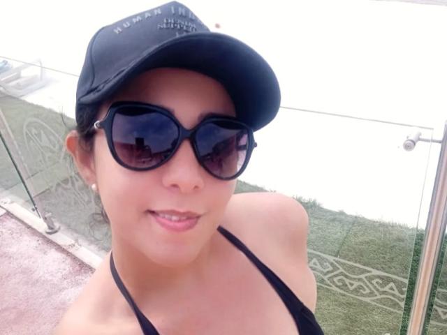 Katty Rivera, con gorra negra, gafas de sol negras y top halter negro, sonriendo a la cámara, mostrando los hombros desnudos y un ligero escote, de pie sobre una superficie verde texturizada..