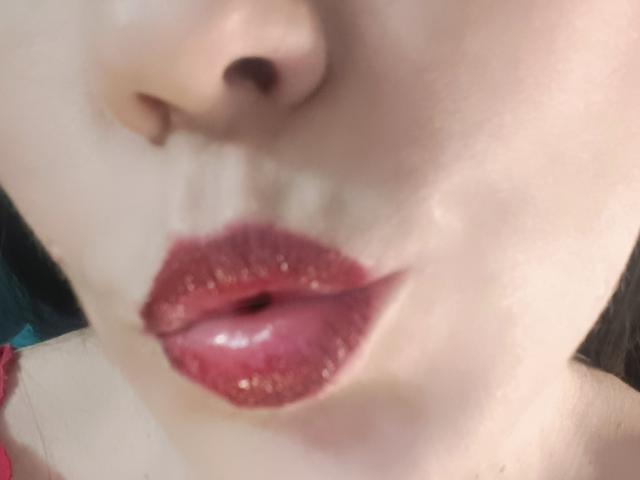 Das Gesicht von OrianaLaFrancaise, wobei der Fokus auf ihren glänzenden, rosa-roten, leicht geöffneten Lippen liegt, mit einem Hauch ihrer Nase und ihrer oberen Wange. Ihre Haut ist hell und glatt..