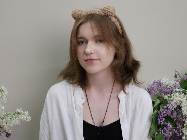 YukiKendal has light skin, brown wavy hair, and wears a beige bear headband. She's in a white shirt over a black top, with lilac and white flowers.