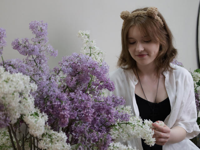 YukiKendal with brown hair, wearing a bear headband and white shirt, holding purple and white lilacs, smiling softly, black dress underneath, standing among abundant flowers.