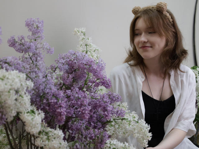YukiKendal with brown hair, cat ears headband, white shirt, black top, smiling at purple and white lilac flowers. Her small tits are slightly visible under her shirt.