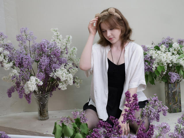 YukiKendal with light brown hair, wearing a beige headband, white shirt, black dress, sitting among purple and white lilacs, touching her head. She has a cute, shy expression.