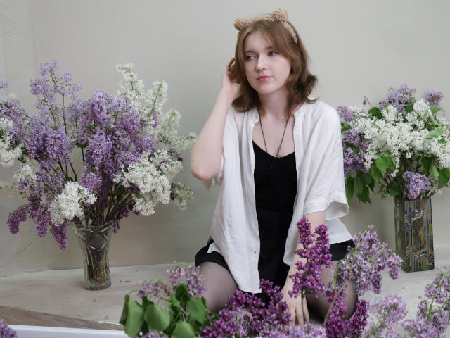 YukiKendal with wavy brown hair, wearing a white shirt over a black dress, sitting among purple and white lilacs, wearing a cute bear headband, looking slightly bored.
