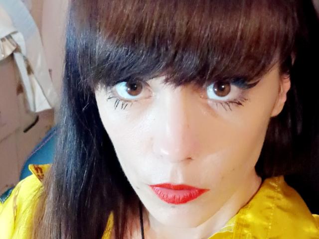 OrianaLaFrancaise has dark brown eyes, red lipstick, and straight brown bangs covering her forehead. She's wearing a yellow shirt with a shiny texture. Her makeup is dramatic.