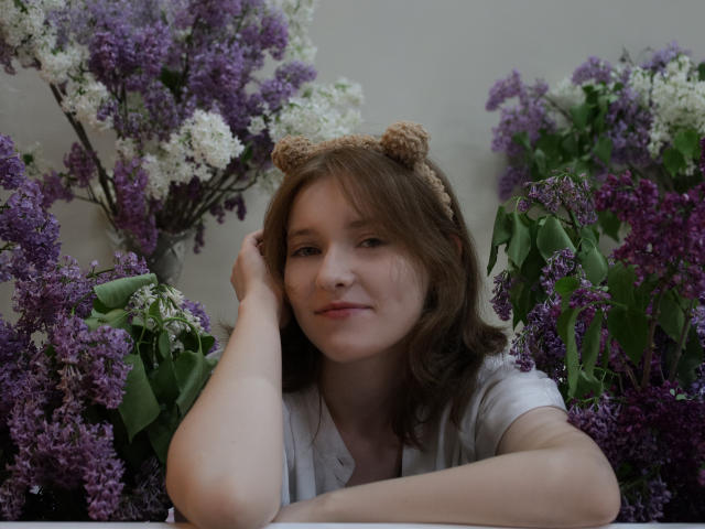 YukiKendal with brown hair, cute bear headband, white shirt, arms crossed, surrounded by purple and white lilacs, slight smile, relaxed, gentle expression, soft lighting, natural setting.