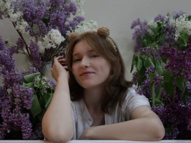 YukiKendal with brown hair, wearing a white shirt, cute bear headband, smiling, surrounded by purple and white lilacs, resting her chin on her hand, looking relaxed.