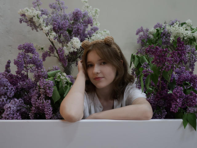 YukiKendal with brown hair, wearing a white shirt and bear headband, leaning on a white wall, surrounded by purple and white lilacs, looking relaxed and cute.