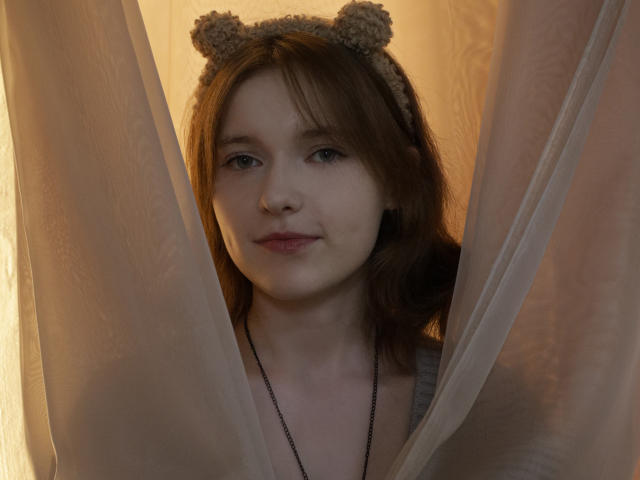 YukiKendal with brown hair, wearing a fuzzy bear headband, looking through sheer curtains, wearing a gray tank top and black necklace, has a cute, soft expression.