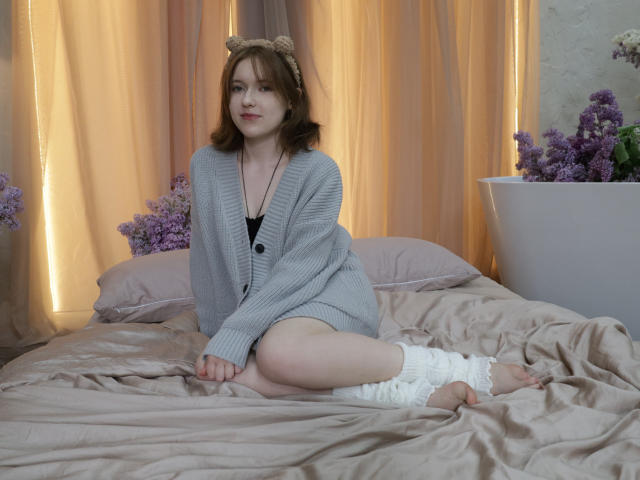 YukiKendal with medium tits, wearing a gray cardigan, black bra, and white leg warmers, sits on a bed with beige sheets, brown hair, cat ears, and purple flowers.