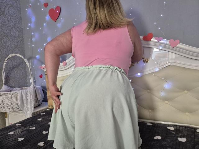 RadaRori with blonde hair, pink top, and green skirt, facing away, showing her ass, standing in front of a white, tufted bed with heart decorations and fairy lights.