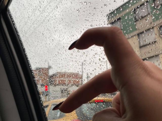AmaraKazumis Hand mit schwarz lackierten Nägeln berührt eine von Regentropfen bedeckte Autoscheibe, wobei feuchte Finger vor einem grauen, bewölkten Hintergrund zu sehen sind. Ein gelbes Taxi ist draußen sichtbar..