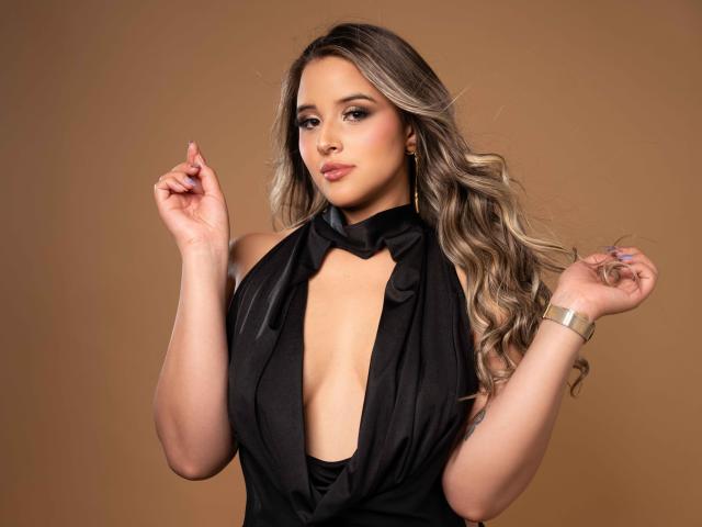 SunnyLuxx has long, wavy blonde hair, wearing a black, deep-cut dress, exposing her tits, with gold earrings and a gold bracelet, against a brown background.